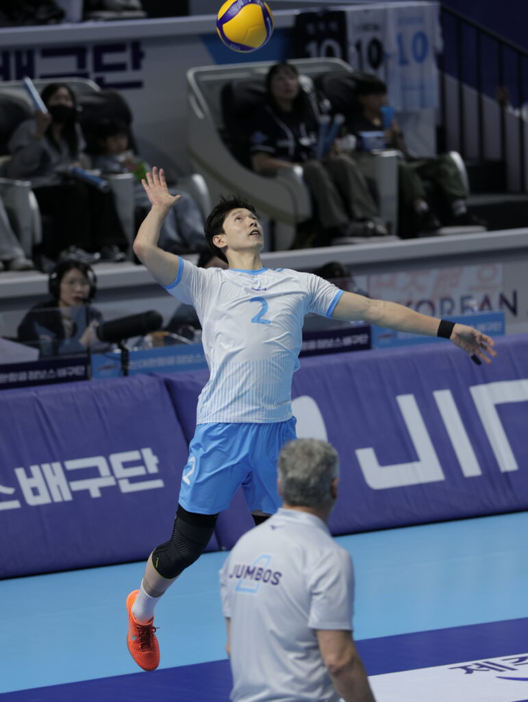 Volleyball player serving during a match in an indoor arena.